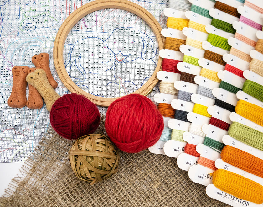 Embroidery hoop with cross-stitch pattern, red yarn, wooden beads, and thread spools on a textured surface.