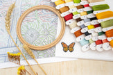 Embroidery hoop with cross-stitch pattern, colorful threads, and butterfly decoration on a table.