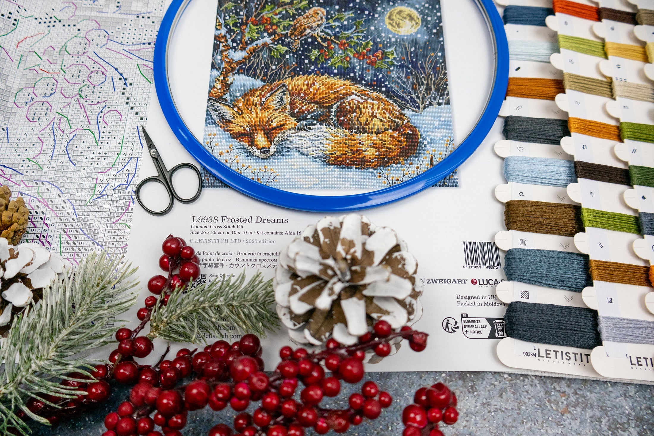Embroidery hoop with a fox design, scissors, thread spools, and decorative items on a table.