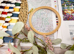Embroidery hoop with cross-stitch pattern, threads, and decorative elements on a wooden surface.