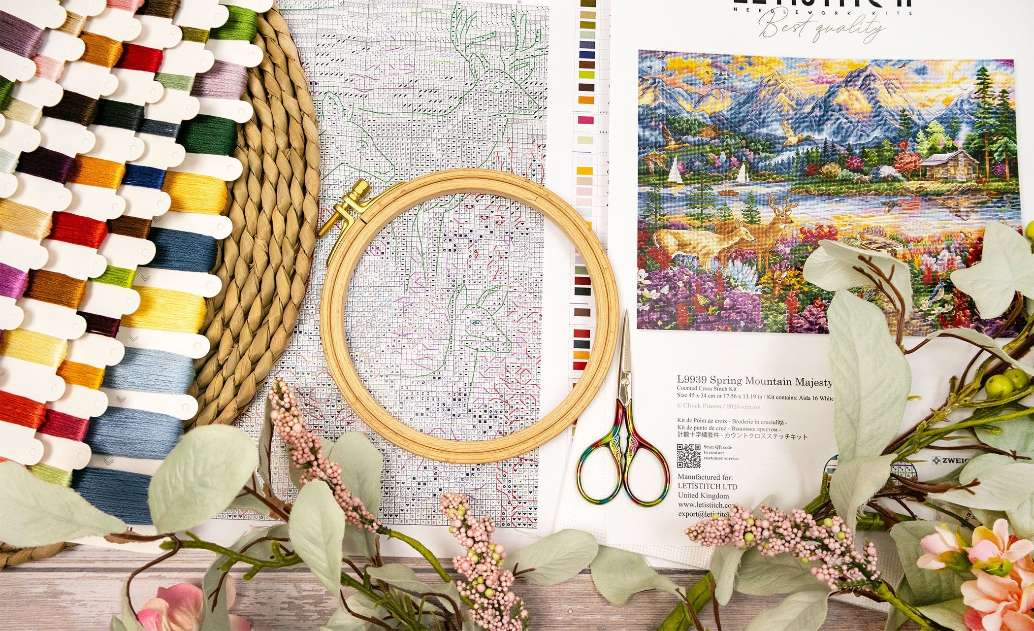 Embroidery hoop with colorful threads, scissors, and a scenic painting on a wooden surface.