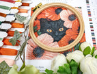Embroidery hoop with a black cat surrounded by flowers on a floral-patterned fabric background.