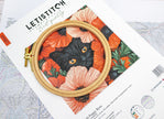 Embroidery hoop with a black cat design on floral fabric, placed on a Letistitch embroidery kit packaging.