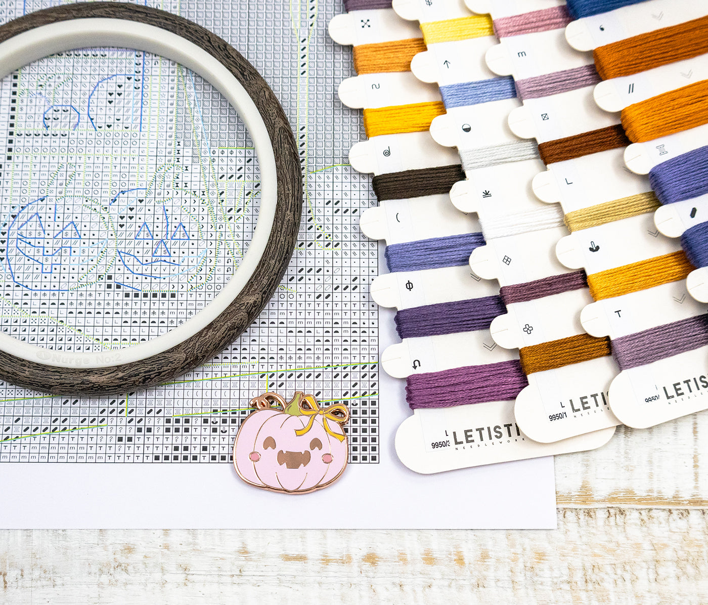 Embroidery hoop with cross-stitching design, colorful threads, and a small pumpkin charm on a wooden surface.