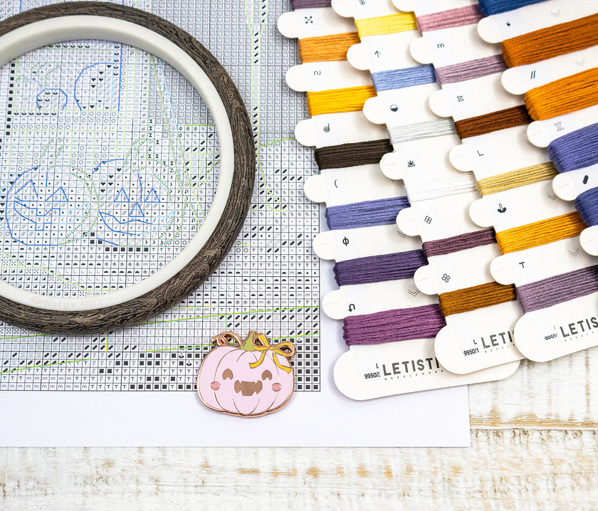 Embroidery hoop with cross-stitching design, colorful threads, and a small pumpkin charm on a wooden surface.