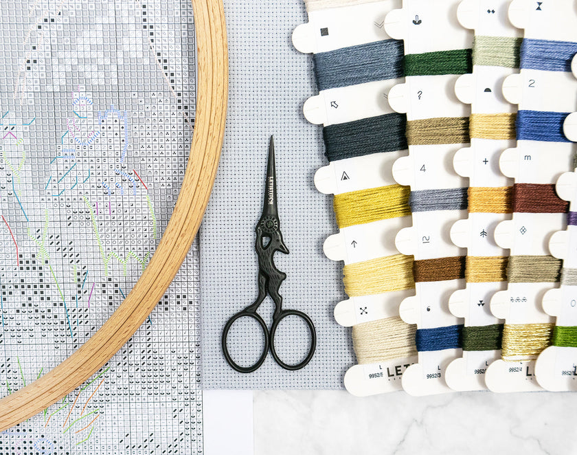 Embroidery hoop, scissors, and thread spools on a cross-stitching fabric.