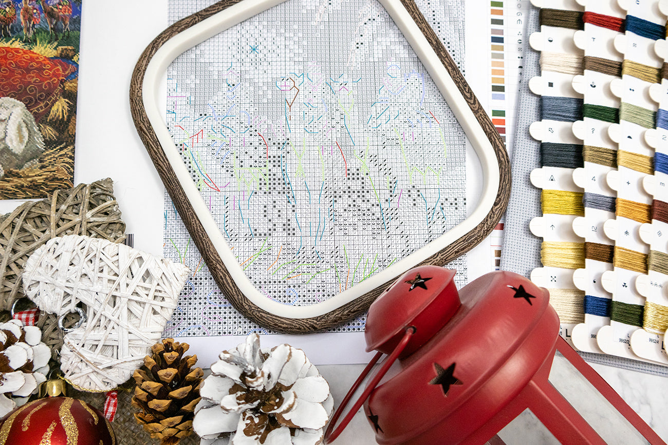 Cross-stitching hoop with partially completed design, red lantern, pinecones, and thread spools on a table.