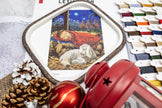 Embroidery hoop with nativity scene design, red bell, pine cones, and Christmas decorations on a white surface.