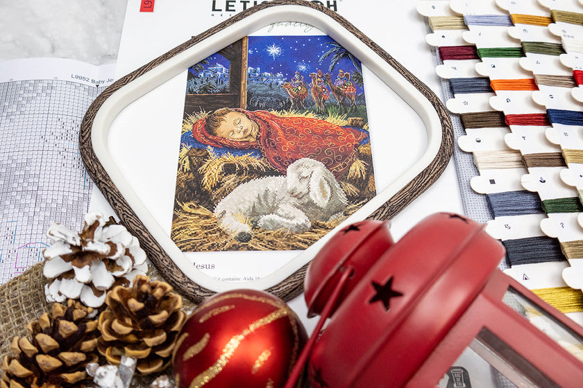 Embroidery hoop with nativity scene design, red bell, pine cones, and Christmas decorations on a white surface.