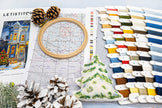 Cross-stitching materials including an embroidery hoop, thread colors, and a Christmas-themed fabric swatch on a light surface.