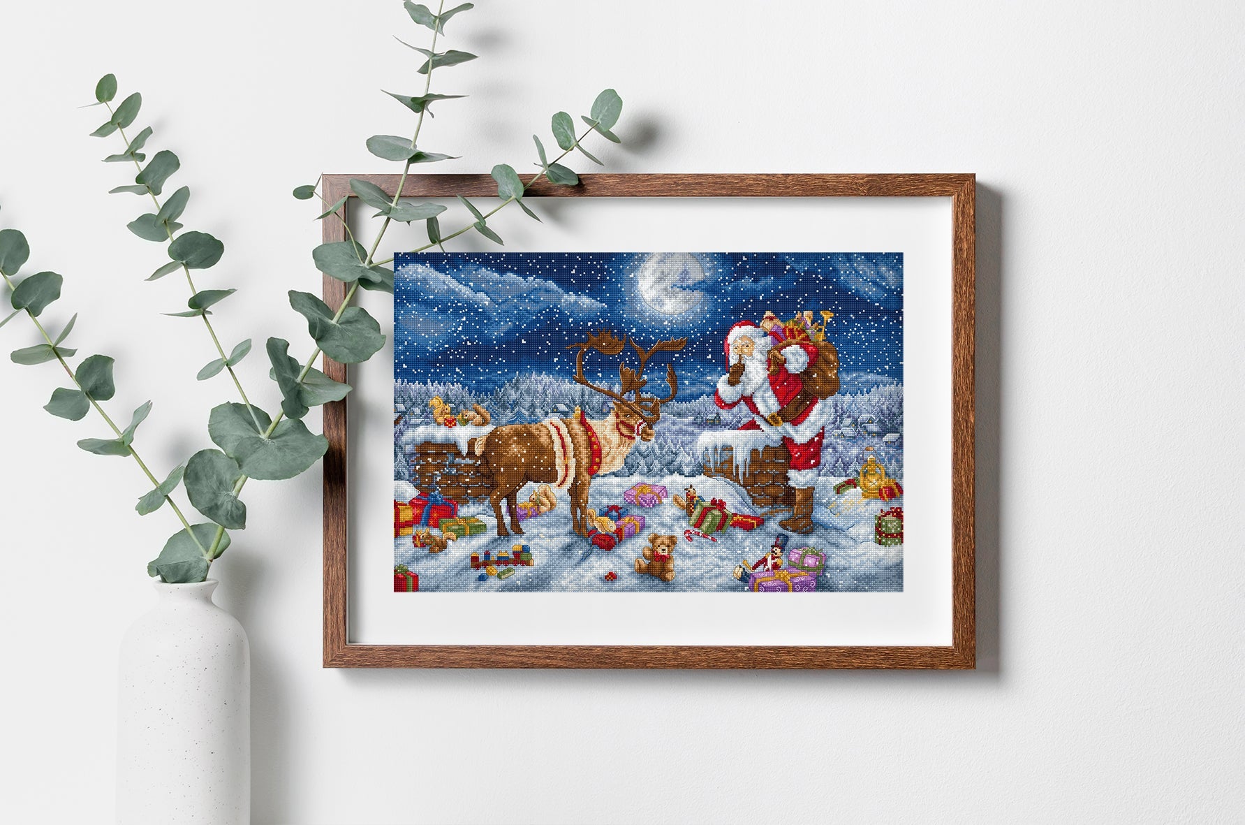 Framed artwork of Santa Claus and reindeer with Christmas scene, surrounded by eucalyptus leaves.