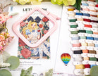 Cross-stitch kit with design, thread spools, and decorative elements on a magazine cover.