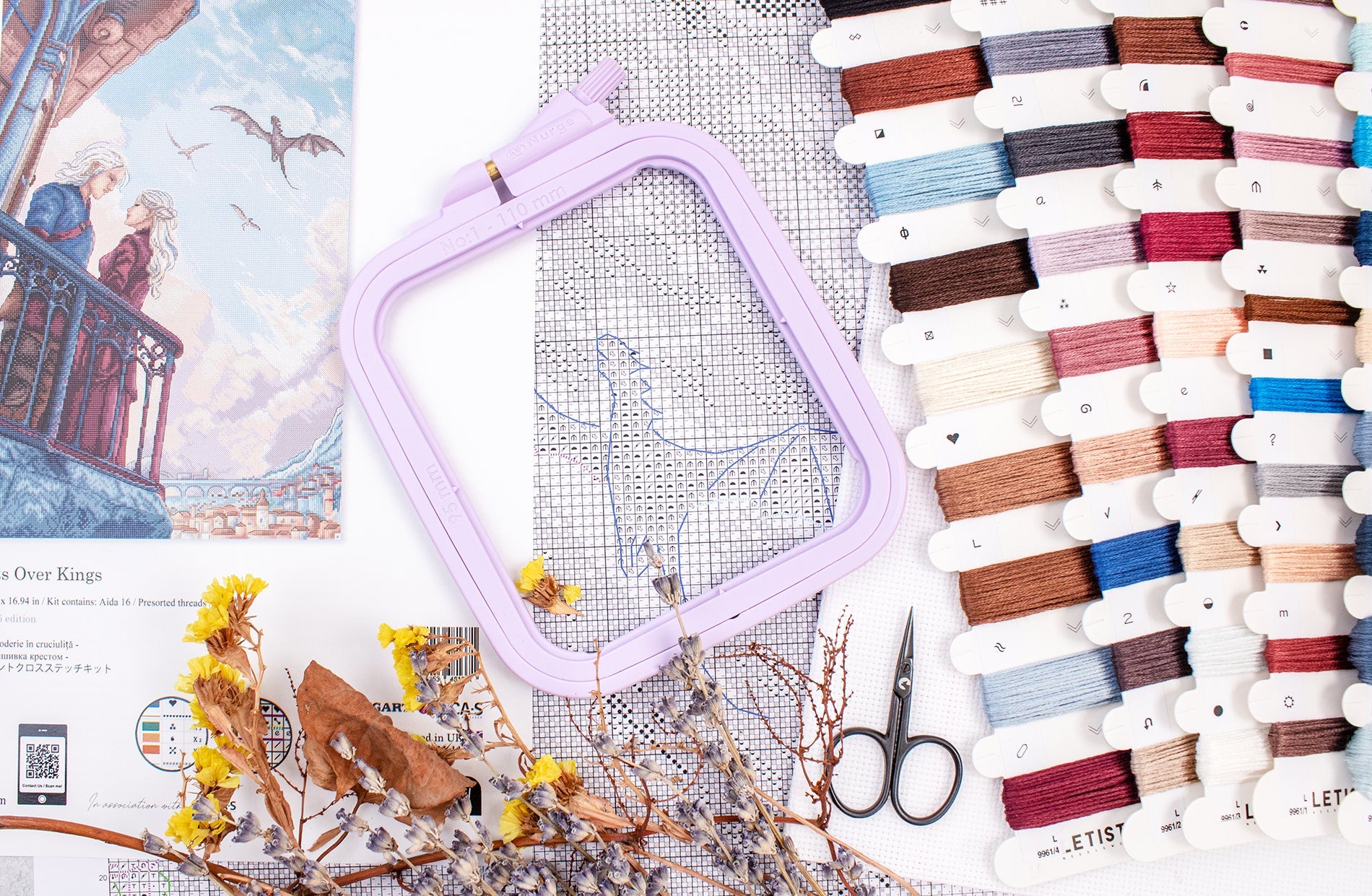 Embroidery hoop, cross-stitch pattern, scissors, and spools of thread on a surface.