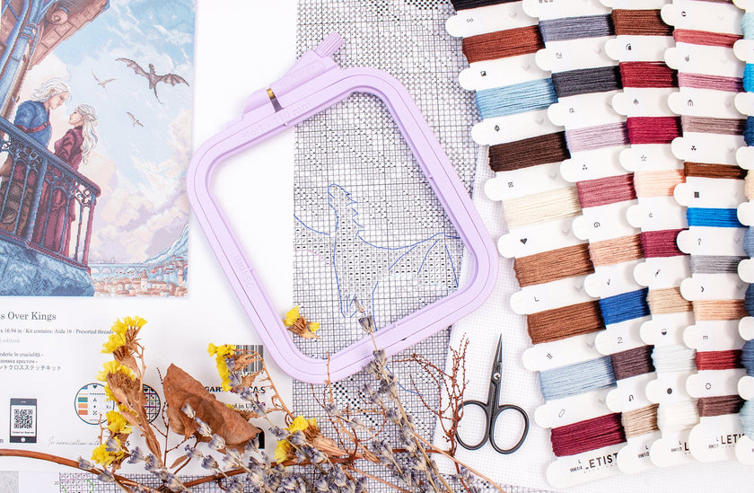 Embroidery hoop, cross-stitch pattern, scissors, and spools of thread on a surface.