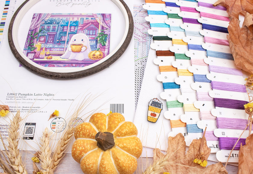 Embroidery hoop with a pumpkin-themed design, colorful thread spools, and pumpkins on a table.