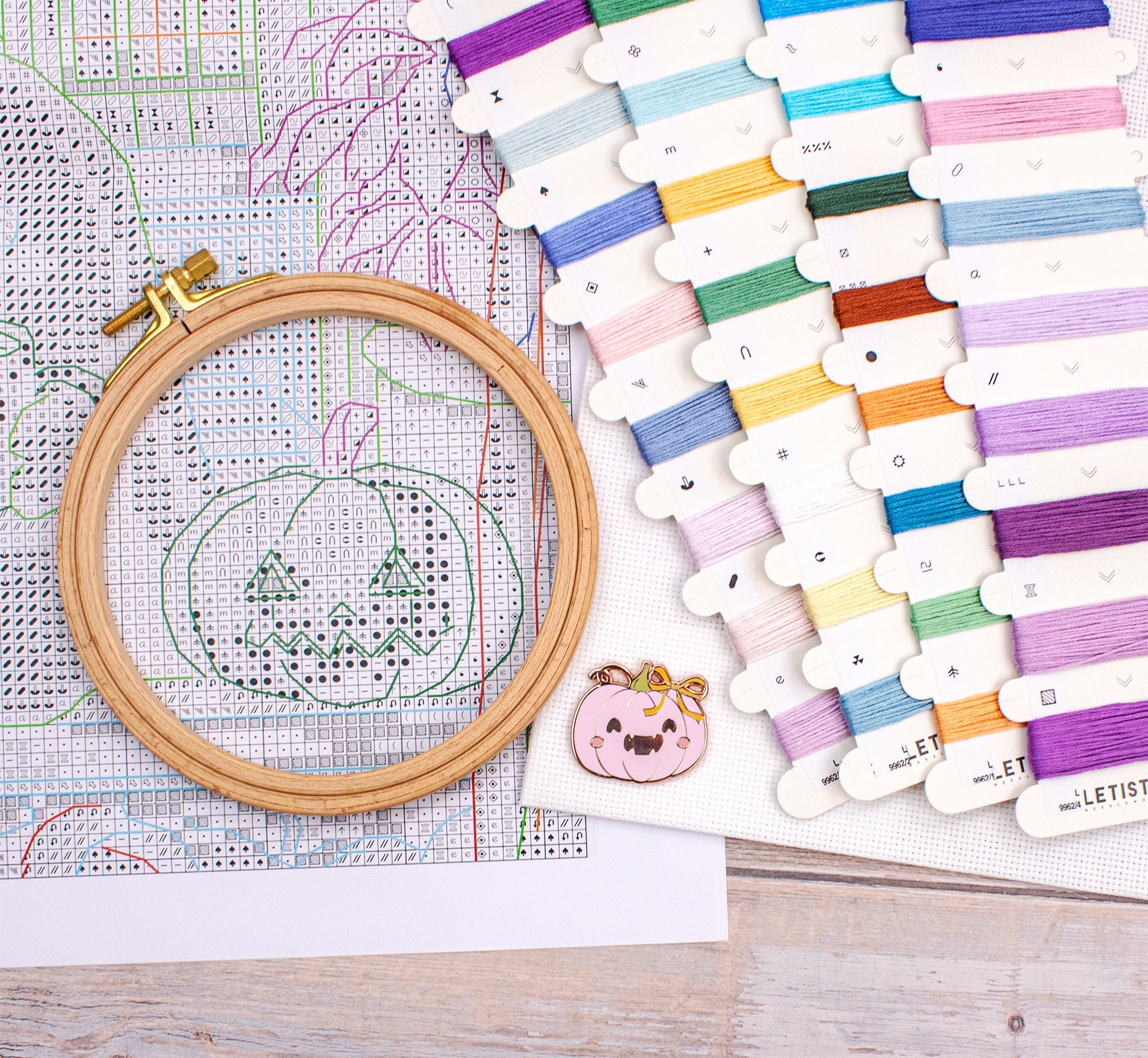Embroidery hoop with pumpkin design on cross-stitch pattern, surrounded by colorful threads.