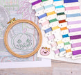 Embroidery hoop with pumpkin design on cross-stitch pattern, surrounded by colorful threads.