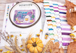 Embroidery hoop with a colorful design, surrounded by threads and autumn decorations on a wooden surface.