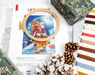 Cross-stitch kit with Santa Claus design, embroidery hoop, and Christmas-themed packaging on a white surface.