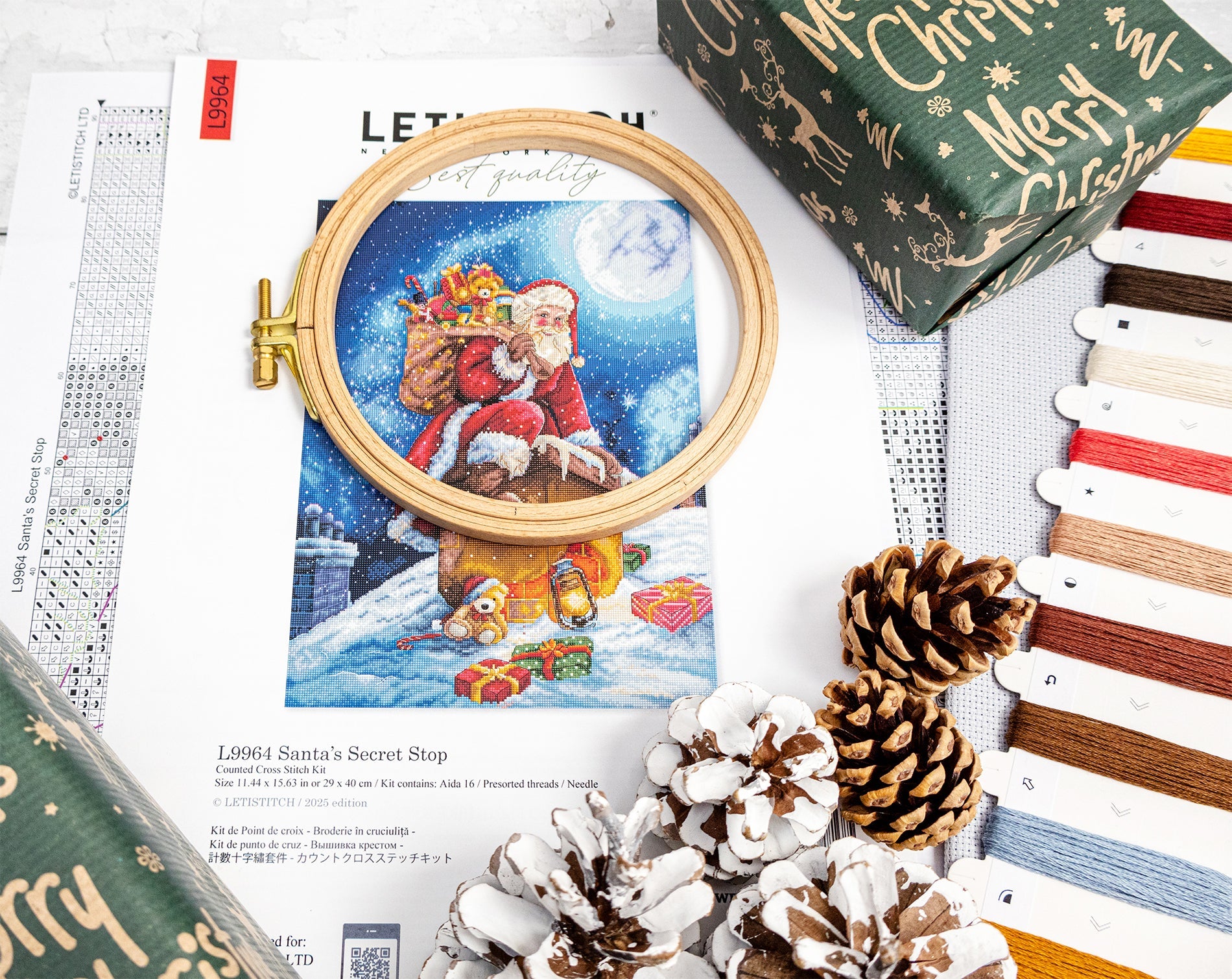 Cross-stitch kit with Santa Claus design, embroidery hoop, and Christmas-themed packaging on a white surface.