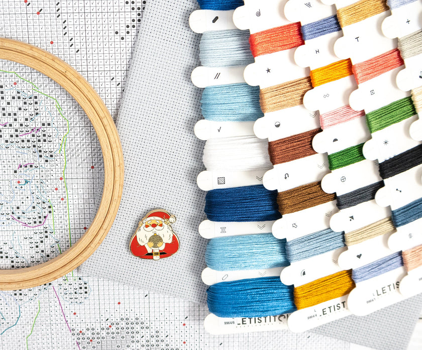 Embroidery hoop with cross-stitch pattern and color swatch of embroidery threads on a textured fabric background.