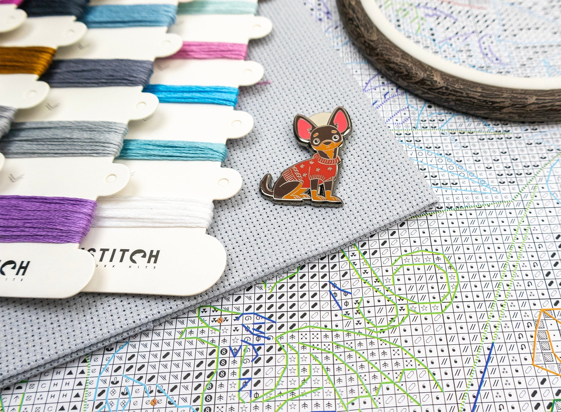 Dog-themed enamel pin on a cross-stitching fabric with embroidery threads and hoop in the background.