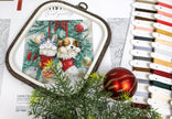 Embroidery hoop with a Christmas-themed cross-stitch design featuring a dog and rabbit in red booties, surrounded by decorative ribbons and greenery.