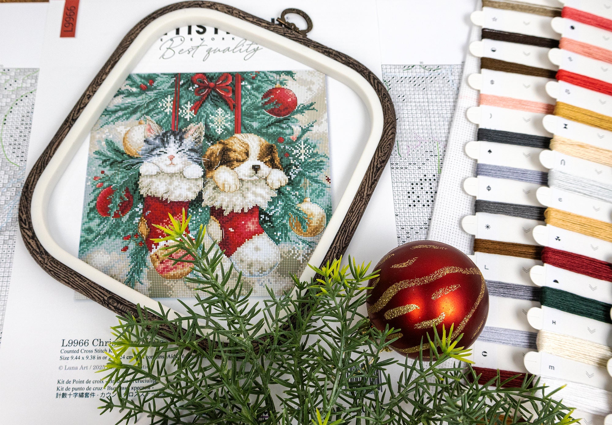 Embroidery hoop with a Christmas-themed cross-stitch design featuring a dog and rabbit in red booties, surrounded by decorative ribbons and greenery.