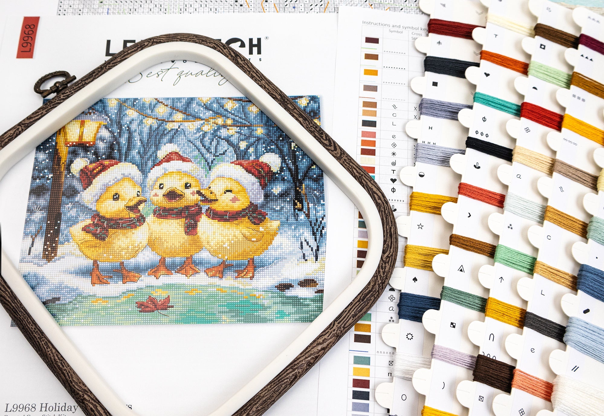 Cross-stitch kit with three yellow chicks in winter hats and scarves, surrounded by embroidery threads and color swatches.