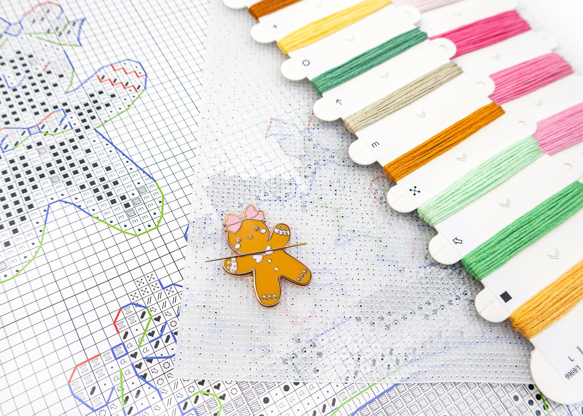 Colorful embroidery floss spools with a gingerbread man pin on a cross-stitch pattern.