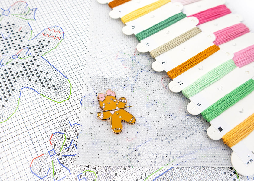 Colorful embroidery floss spools with a gingerbread man pin on a cross-stitch pattern.