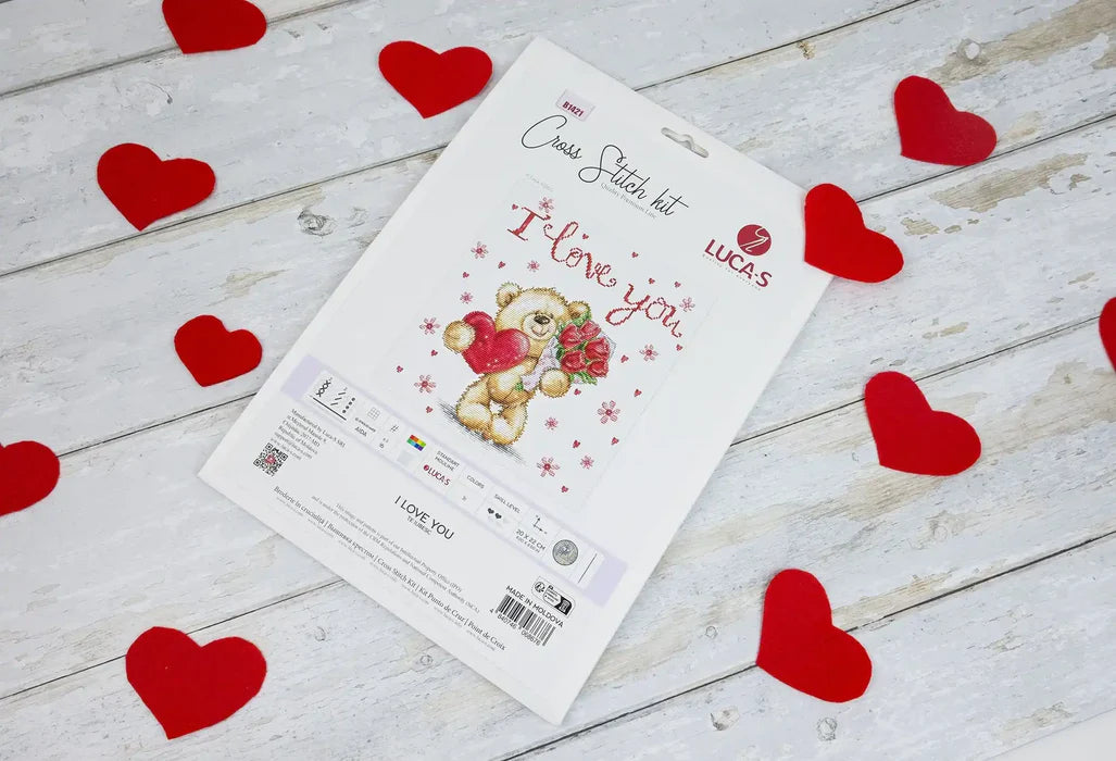 Cross stitch kit with a teddy bear design surrounded by red hearts on a wooden surface