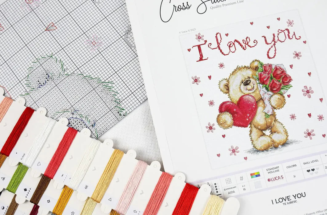 Cross stitch kit with a teddy bear design and 'I love you' text, surrounded by threads and a cross stitch chart.