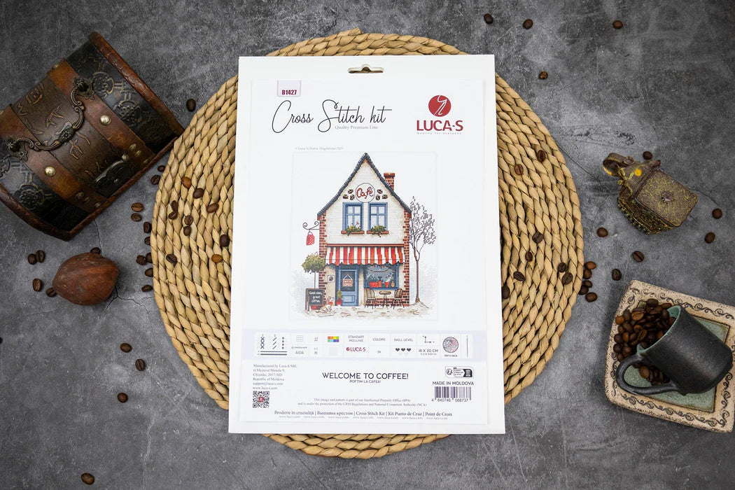 Cross stitch kit packaging on a textured surface with coffee beans and a mug.