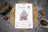 Cross stitch kit packaging on a textured surface with coffee beans and a mug.