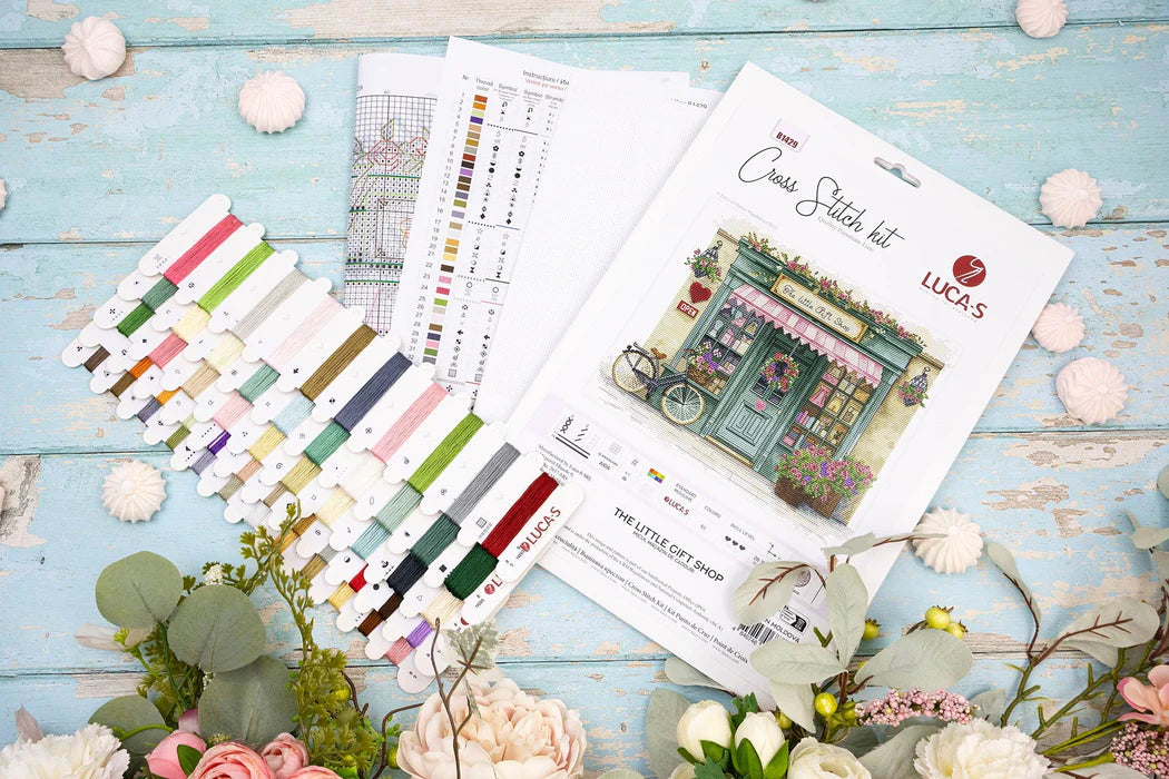 Luca's Cross Stitch kit with threads and pattern sheets on a wooden surface with flowers.