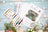 Luca's Cross Stitch kit with threads and pattern sheets on a wooden surface with flowers.