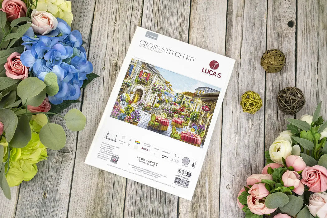 Cross-stitch kit packaging on a wooden surface with flowers around