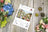 Cross-stitch kit packaging on a wooden surface with flowers around
