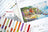 Color chart and thread spools with a scenic painting of a village by a lake, featuring the brand 'Lucas'.