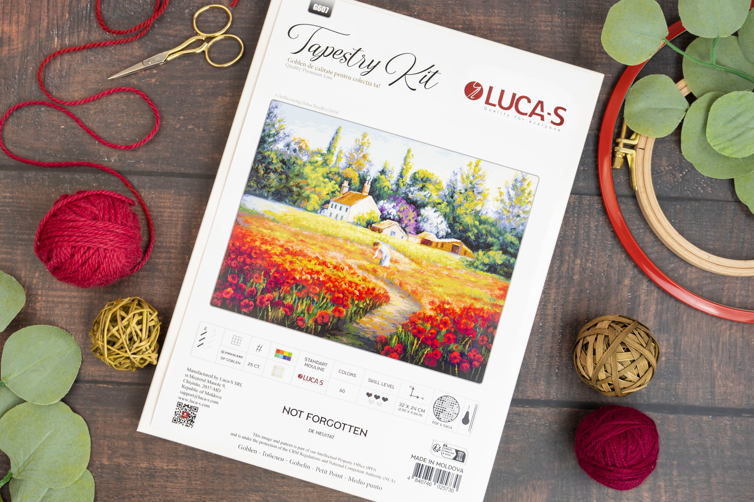 Luca's Tapestry Kit needlepoint canvas shown in a styled flat-lay on a dark wood background, surrounded by craft supplies including red yarn balls, gold scissors, a wooden hoop, and green eucalyptus leaves. The canvas features a colorful landscape painting of a field of red and orange flowers with a small white house.
