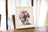 Framed artwork of a floral arrangement on a wooden table with white curtains in the background.
