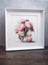 Framed artwork of pink flowers in a glass vase on a wooden surface with a gray background