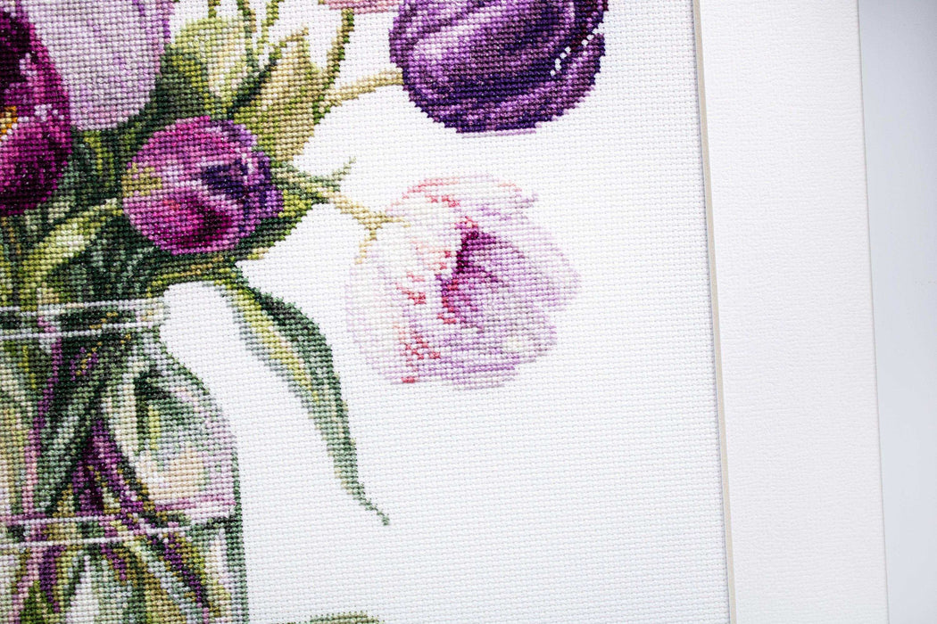 Floral cross-stitch pattern with purple and pink flowers in a glass jar on white fabric.