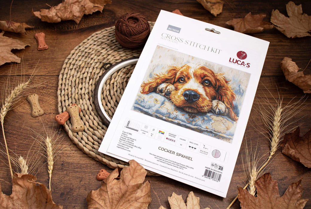 Cross-stitch kit for a Cocker Spaniel on a wooden surface with fall leaves and a woven mat.