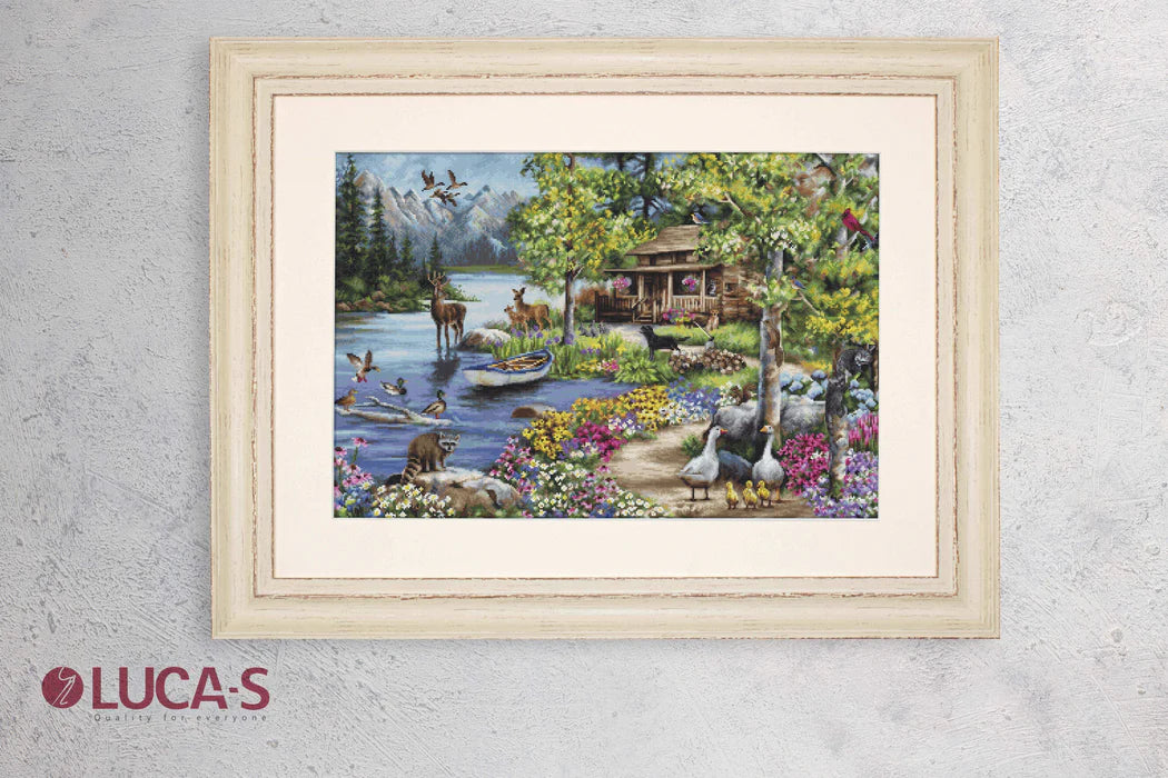 Framed cross-stitch artwork of a lakeside scene with animals and flowers, branded 'Lucas'.