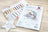 Cross stitch kit with a design of an elephant on a moon, surrounded by color swatches and branding.