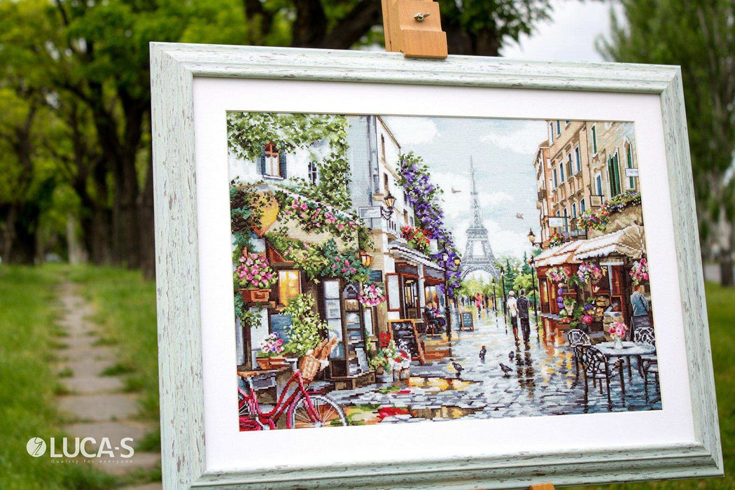 Framed artwork of a street scene with cafes and people, displayed outdoors.