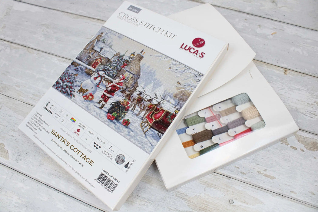 Cross stitch kit packaging with Santa's Cottage design on a wooden surface