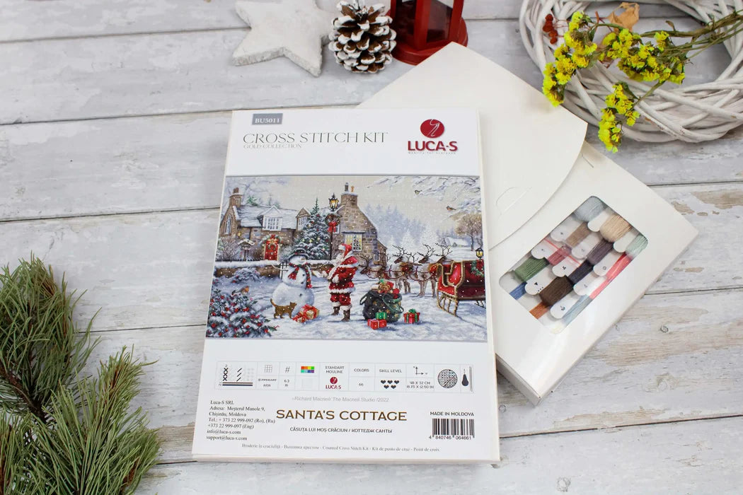 Lucas cross-stitch kit for 'Santa's Cottage' on a wooden surface with Christmas decorations.